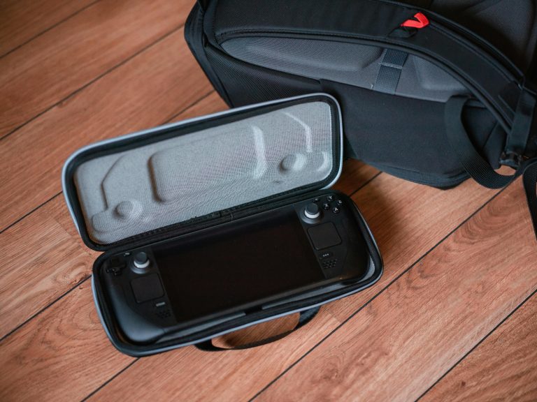 A portable gaming console in a protective case on wooden floor with backpack.