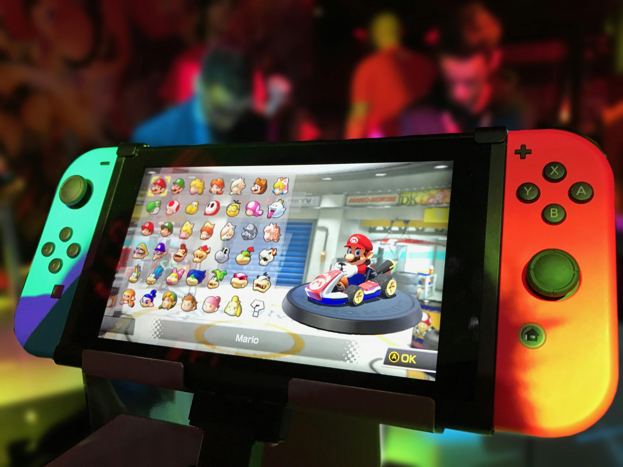 Close-up of a Nintendo Switch showing Mario Kart selection screen, gaming atmosphere in the background.